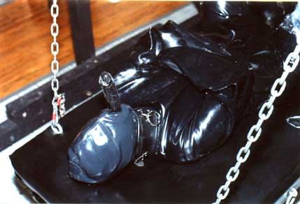 Dominatrix Sharina Nicole has strapped her submissive in a latex cat suit, latex straight jacket and fitted him with a fetish latex hood complete with rubber penis attachment.